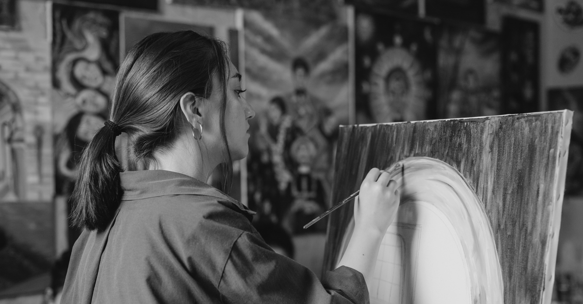 A woman artist paints on a canvas in a studio filled with artwork, captured in black and white.